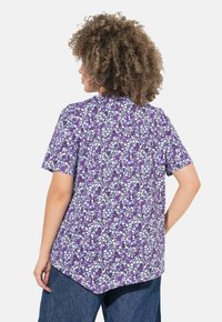 Maglietta viola a maniche corte con motivo a mosaico e accenti bianchi e viola scuro. L'orlo è più lungo sul retro ed è realizzata in materiale morbido.