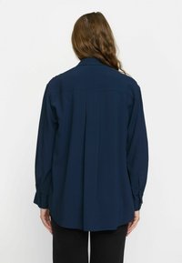 Chemise bleu marine à boutons, à manches longues, avec un pli au dos et un ourlet arrondi, fabriquée en tissu lisse. Un pantalon noir est porté.