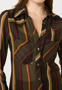 Chemise boutonnée à rayures en tissu transparent, présentant des rayures verticales en vert foncé, marron, jaune et rose clair. Comprend deux poches poitrine.