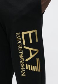 Fekete melegítőnadrág sima textúrával, arany "EA" és "EMPORIO ARMANI" logókkal az oldalon, barna részletekkel a derékpánton.