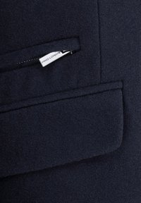 Granatowa kurtka z wełny z dodatkiem, z kieszenią na zamek oraz metalowymi elementami, podszewką i wyraźnie zarysowanym kołnierzem, o zarysowanej konstrukcji i miękkiej teksturze.