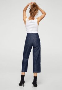Jeans de mezclilla azul de cintura alta con un diseño de pierna ancha recortada, que cuentan con un dobladillo deshilachado y dos bolsillos traseros. Se llevan con una camiseta sin mangas blanca.