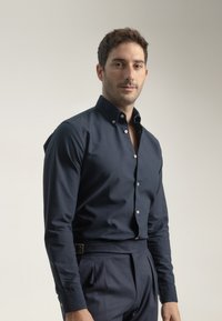 Camisa de botón en color azul marino con un cuello estructurado, mangas largas y botones blancos, combinada con pantalones oscuros a medida. Tela suave con un ligero brillo.