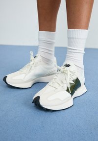 Balti sportiniai bateliai su lygiu paviršiumi, turintys blizgų alyvuogių žalios akcentą ir juodą guminį padas. Šnerves pagamintos iš tekstūruotos medžiagos.