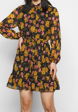 Vestito floreale nero trasparente con fiori gialli e rosa, maniche lunghe, chiusura con bottoni sul davanti, vita annodata e orlo arricciato sopra le ginocchia.