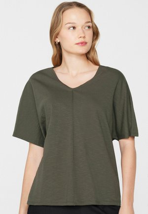 Joven con cabello rubio hasta los hombros, vistiendo una blusa suelta de manga corta y cuello en V color verde oscuro, mirando hacia su derecha.