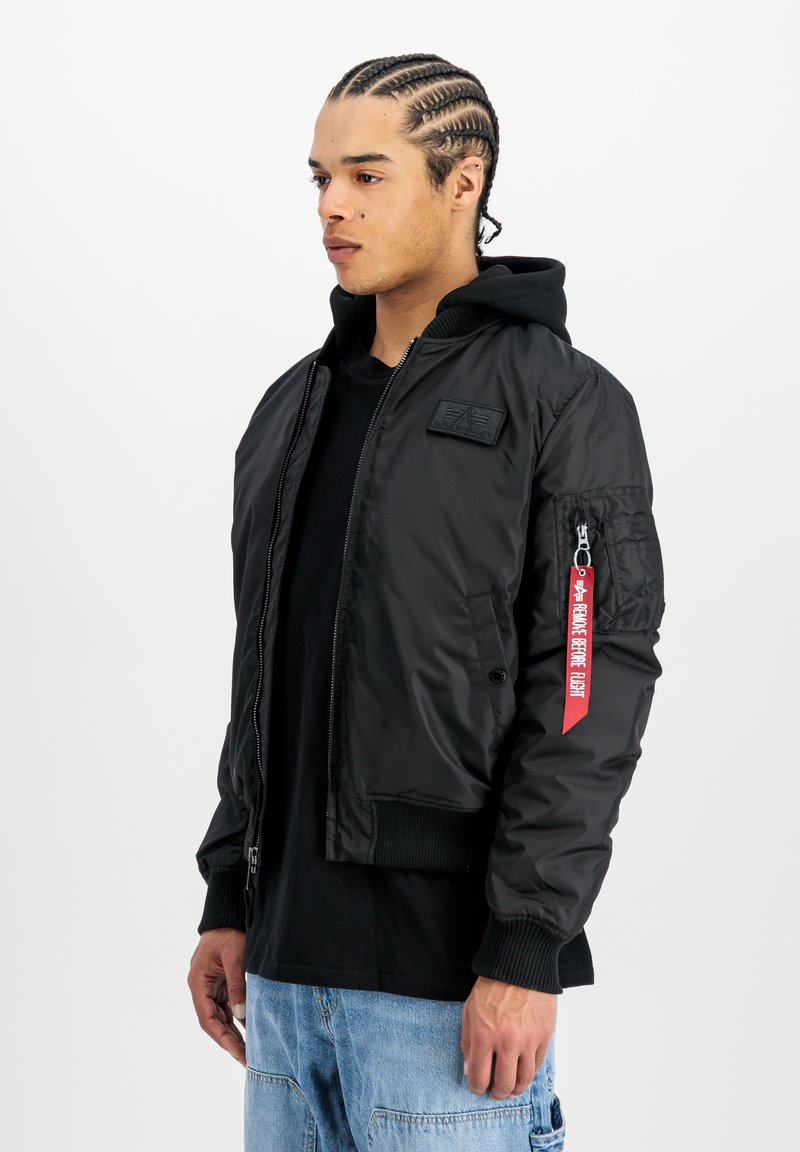 Chaqueta bomber negra con cierre frontal, puños acanalados y cuello. Presenta un parche en el pecho y una etiqueta roja en la manga.