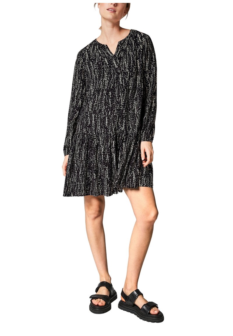 Schwarzes Kleid mit weißem gesprenkeltem Muster, langen Ärmeln, V-Ausschnitt, gerafftem Taillenbund und gestuftem Rock; getragen mit schwarzen klobigen Sandalen.