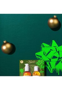 Zielona teksturowana powierzchnia z dwoma złotymi ozdobami. Na pierwszym planie znajduje się karta produktu Kiehl's z dwoma butelkami pompkowymi, jedną w kolorze bursztynowym i jedną przezroczystą.