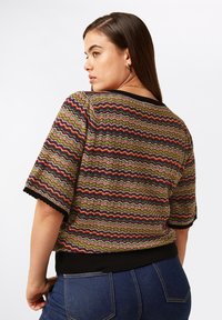 Strickoberteil mit einem Zickzackmuster in grünen, roten und violetten Farbtönen, mit schwarzem Saum und kurzen Ärmeln, kombiniert mit dunkelblauen Jeans.