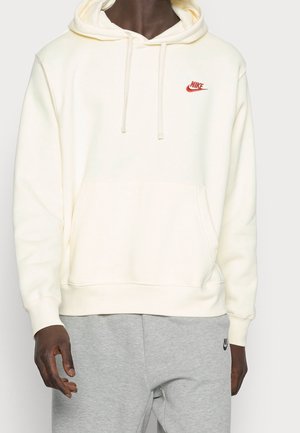 Osoba nosząca kremową bluzę z kapturem Nike z przednią kieszenią i sznurkami, w połączeniu z jasnoszarymi spodniami dresowymi Nike.