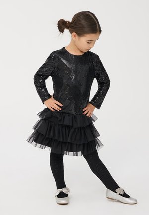 Top di paillettes nere con una gonna di tulle a volant, abbinato a collant neri testurizzati e ballerine argento con un accento di fiocco.