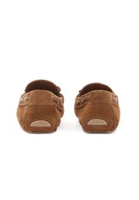 Καφέ σουέντ μοκασίνια φαίνονται από τη φτέρνα, με σχεδιασμό slip-on και λαστιχένιες επιφάνειες στη φτέρνα, σε λευκό φόντο.