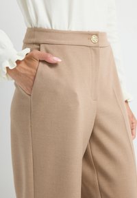 Hand in der Tasche einer hoch taillierten beigen Hose mit goldenem Knopf, kombiniert mit einem weißen Oberteil mit langen Ärmeln und Rüschensaum.