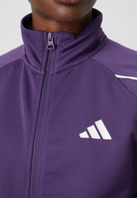 Violet sportinė striukė su aukštu apykakle, užtrauktuku ir baltu prekės ženklu. Lygus paviršius su kontrastuojančiomis baltomis detalėmis ant pečių.