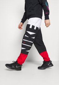 Person läuft in schwarzen, weißen und roten Nike-Hosen mit großem Logo und schwarzen Nike-Sneakern vor einem einfarbigen Hintergrund.