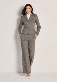 Vrouw staat met handen in de zakken, gekleed in een bruine gestreepte pak, witte blouse, opvallende oorbellen en witte puntige hakken.