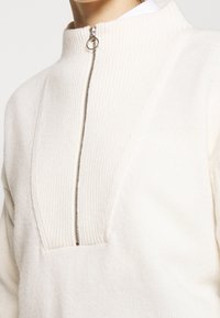 Pull en maille blanche avec un col montant, fermeture zippée mi-hauteur à l'avant, et texture côtelée avec un design en panneaux prononcés.
