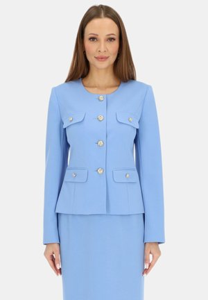 Kobieta o prostych, brązowych włosach nosi dopasowaną jasnoniebieską marynarkę z złotymi guzikami oraz pasującą spódnicę, stojąc na jednolitym tle.