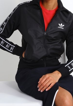 Osoba ubrana w czarną kurtkę sportową Adidas z białym logo i paskami, siedząca z jedną ręką w kieszeni, a drugą opartą na kolanie.