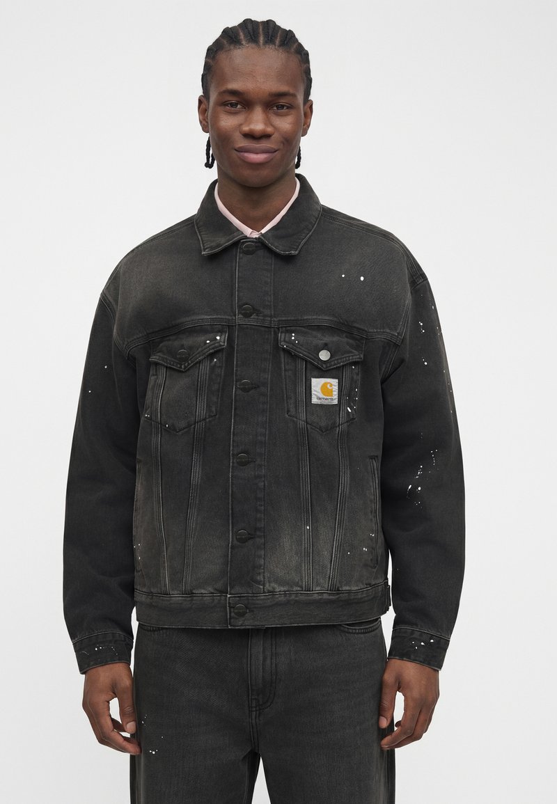 Young man with braided hair wearing a black denim jacket with white paint splatters and matching black pants against a plain background.