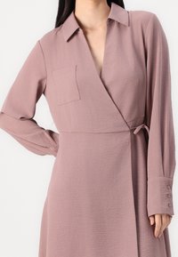 Dlouhé rukávy, zavinovací šaty ve měkké mauve tkanině. Mají límec, náprsní kapsu a zavazování v pase. Rukávy končí na knoflíkové manžetě.