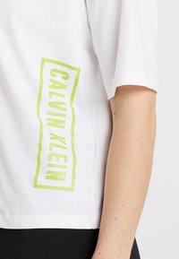 Camiseta de algodón blanca con mangas cortas, que presenta un logotipo vertical en neón verde de "Calvin Klein" en el lado izquierdo. Textura suave.