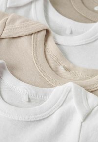 Stapel von Babybodys in neutralen Farbtönen, Nahaufnahme von gerippten Halsausschnitten und Schulternähten in Beige-, Weiß- und Cremetönen.