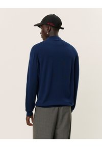 Egy férfi sötétkék pulóverben, szürke nadrágban, piros hímzéses fekete sapkával és gyöngyfülbevalóval, hátulnézetből, világos háttér előtt.