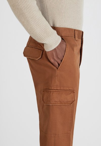 Bruine cargobroek van katoenen stof met zijkanten cargozakken en een aangesloten pasvorm. Een crème ribgebreide trui wordt gedragen, deels zichtbaar.