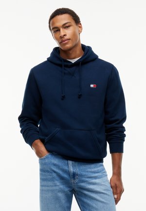 Unge mand iført en mørkeblå hættetrøje med et lille logo og lyseblå jeans, der står mod en ensfarvet hvid baggrund.