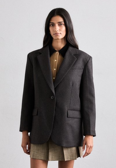 Kulgrå oversized blazer med notched lapels, enkelt knaplukning og forlommer, båret over en brun skjorte med krave og rutet nederdel.