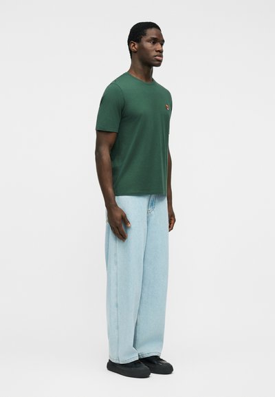 Άνδρας όρθιος, φοράει σκουροπράσινη μπλούζα με κοντά μανίκια και μικρό λογότυπο, ανοιχτόχρωμα χαλαρά τζιν και μαύρα παπούτσια, κοιτάζει προς τα δεξιά.