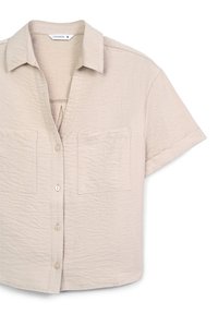 Camicia beige a maniche corte con bottoni realizzata in un tessuto texturizzato, dotata di due tasche frontali e colletto. Progettata con una vestibilità rilassata e cropped.