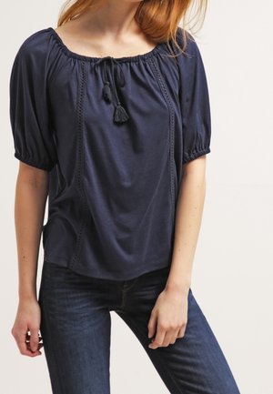 Femme portant un chemisier bleu marine avec des manches courtes bouffantes, un col à nouer avec des pompons, et un jean bleu foncé sur un fond neutre.