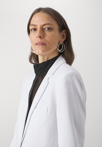 Bijela blazer jakna s odrezanim reverima i jednom džepom, u kombinaciji s crnom bluzom visokog vrata i srebrnim okruglim naušnicama; glatka tekstura.