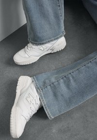 Baskets de sport blanches en cuir texturé avec des accents en maille, accompagnées d'un jean en denim bleu clair, reposant sur une surface grise.