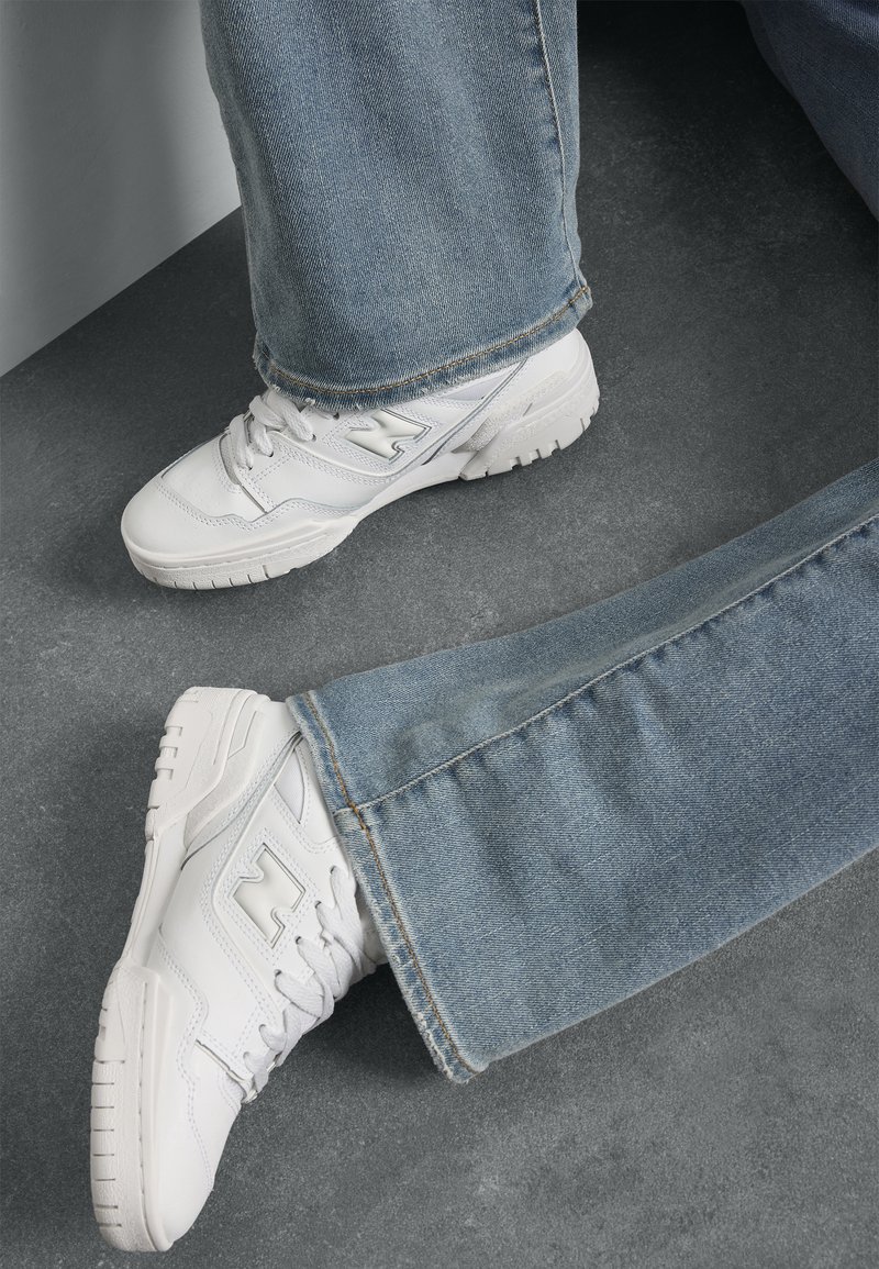 Baskets de sport blanches en cuir texturé avec des accents en maille, accompagnées d'un jean en denim bleu clair, reposant sur une surface grise.