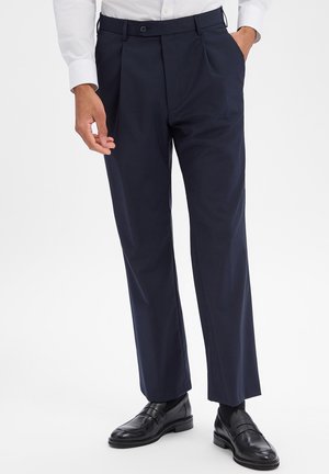 Mann trägt marineblaue, plissierte Anzughose mit schwarzen Leder-Loafers und weißem Hemd, steht mit einer Hand in der Tasche.