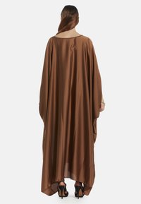 Kaftan in satin marrone con un design ampio e fluente, maniche larghe e scollatura arrotondata, abbinato a scarpe nere con tacco alto.
