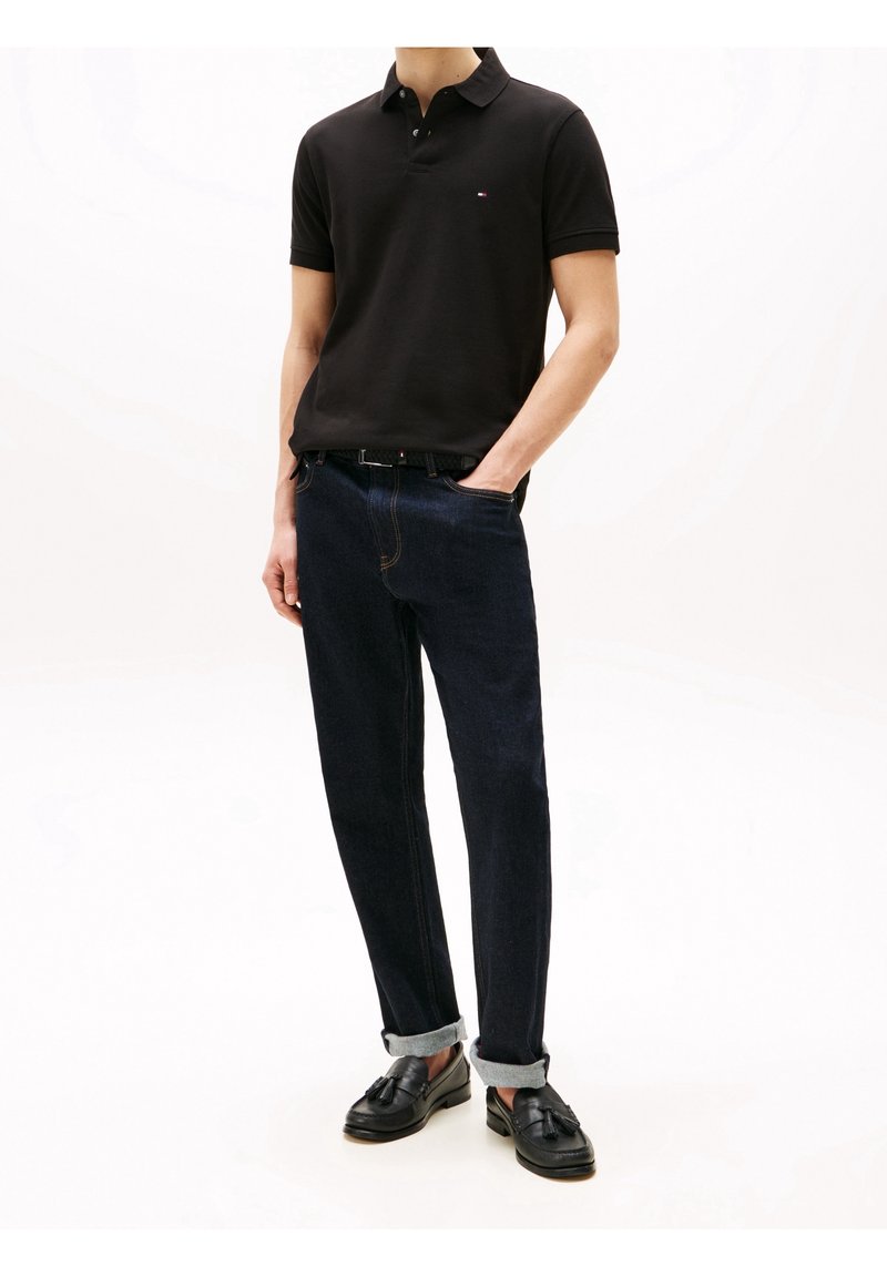 Polo noir, jean en denim avec des poignets clairs et mocassins noirs. Présente un logo subtil sur la poitrine, un design à jambe droite et un délavage foncé.