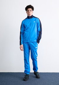 Μπλε αθλητική φόρμα Nike με ζακέτα που έχει φερμουάρ και ναυτικές λεπτομέρειες, συνοδευόμενη από ταιριαστά παντελόνια; μαλακή υφή; φορεμένη με μαύρα αθλητικά παπούτσια.