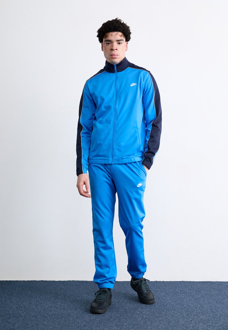 Μπλε αθλητική φόρμα Nike με ζακέτα που έχει φερμουάρ και ναυτικές λεπτομέρειες, συνοδευόμενη από ταιριαστά παντελόνια; μαλακή υφή; φορεμένη με μαύρα αθλητικά παπούτσια.