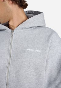 Light gray zip-up hoodie made of soft fabric, featuring a logo on the chest and a spacious hood. Silver zipper adds a contrasting detail.