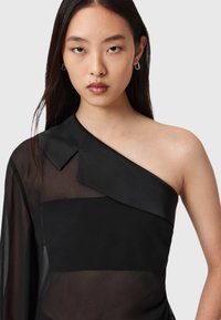 Musta yksi-hartiatoppi, jossa on läpinäkyviä ja kiinteitä paneeleja, terävä kaulus ja sileä pinta, jota täydentävät huomaamattomat korvakorut.