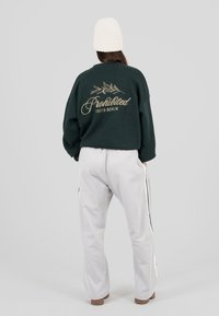 Persona che indossa un berretto lavorato a maglia bianco, una felpa verde scuro con la scritta "Proibito 10119 Berlino" e un design di foglia, e pantaloni grigi chiari con strisce laterali.