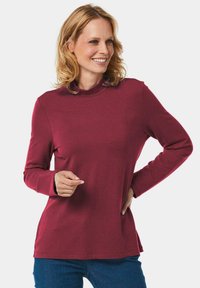 Bordeauxfarbenes Langarmshirt mit hohem Kragen, weicher Textur, taillierter Passform und seitlichen Schlitzern. Kombiniert mit blauen Jeans.