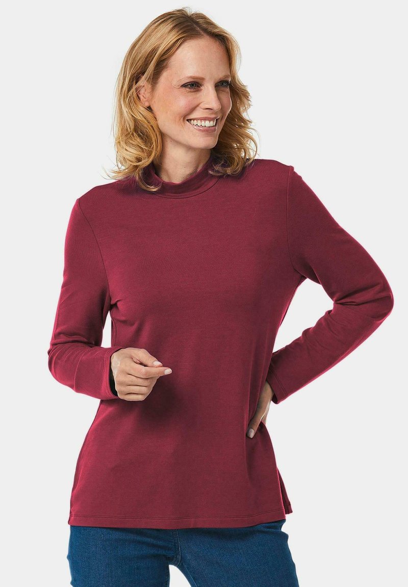 Bordeauxfarbenes Langarmshirt mit hohem Kragen, weicher Textur, taillierter Passform und seitlichen Schlitzern. Kombiniert mit blauen Jeans.