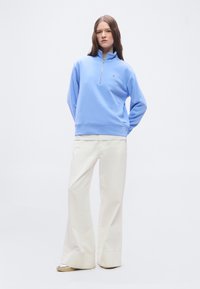 Μια γυναίκα στέκεται φορώντας ανοιχτό μπλε φούτερ με φερμουάρ ως τη μέση, λευκό παντελόνι με φαρδύ πόδι και ασημένια παπούτσια μπροστά σε μια απλή λευκή επιφάνεια.