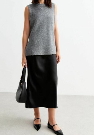 Frau trägt ein ärmelloses graues Stricktop, schwarzen Midirock, schwarze Ballerinas und hält eine schwarze Handtasche an ihrer Seite.